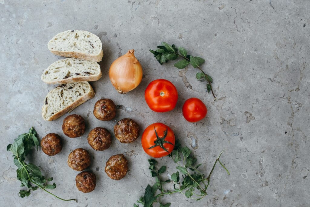 Flat lay of fresh ingredients including bread, meatballs, tomatoes, and herbs on a stone surface.